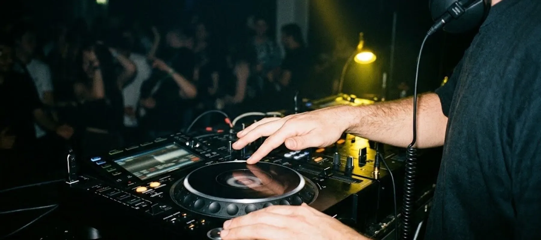 DJ ze słuchawkami koryguje tempo jogiem odtwarzacza, w tle tańczący tłum w żółtym świetle