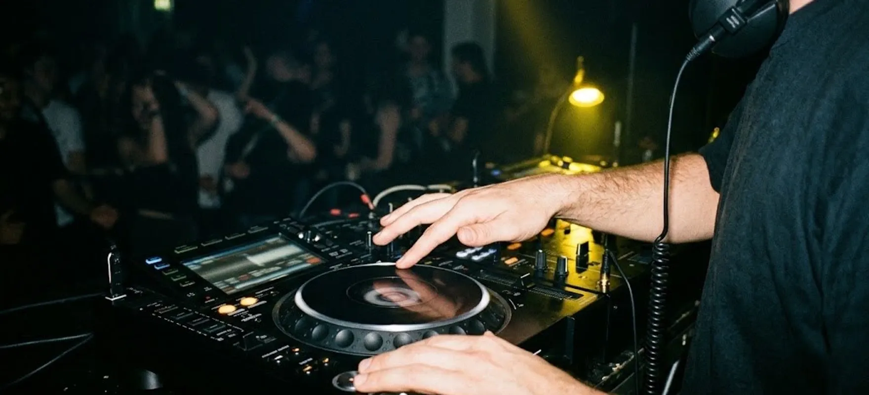 DJ ze słuchawkami koryguje tempo jogiem odtwarzacza, w tle tańczący tłum w żółtym świetle