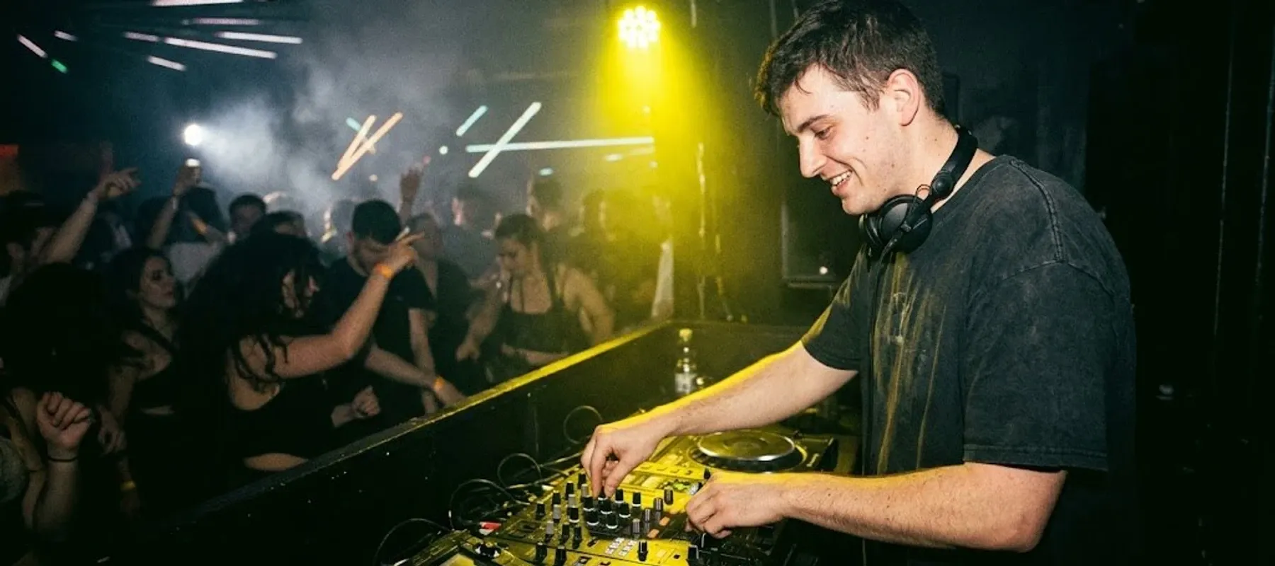 Uśmiechnięty DJ gra swój set przed tańczącą publiką w zadymionym klubie